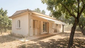 Maison individuelle protégée du soleil estival par des volets extérieurs, un store banne et des arbres feuillus, dans une ambiance réaliste de forte chaleur.