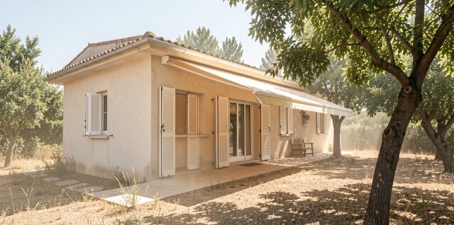 Maison individuelle protégée du soleil estival par des volets extérieurs, un store banne et des arbres feuillus, dans une ambiance réaliste de forte chaleur.