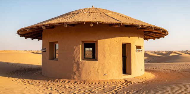 Maison ronde aux murs enduits construits avec des bouteilles plastiques remplies de sable, dotée d’un double toit clair et de petites fenêtres ombragées au milieu d’un désert de dunes dorées.